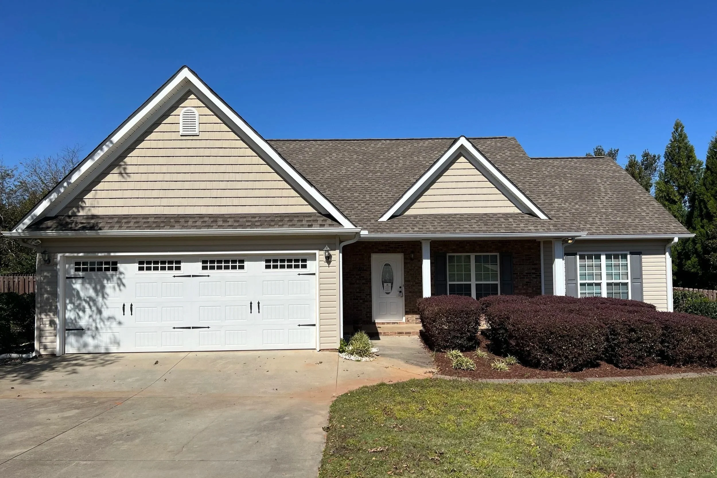 A suburban house with a light beige exterior, attached garage, and a landscaped front yard with bushes and a small lawn, under a clear blue sky.