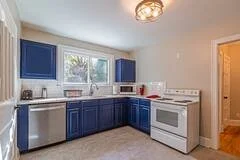 Kitchen with white walls, blue cabinets, a white stove, and a window above the sink.