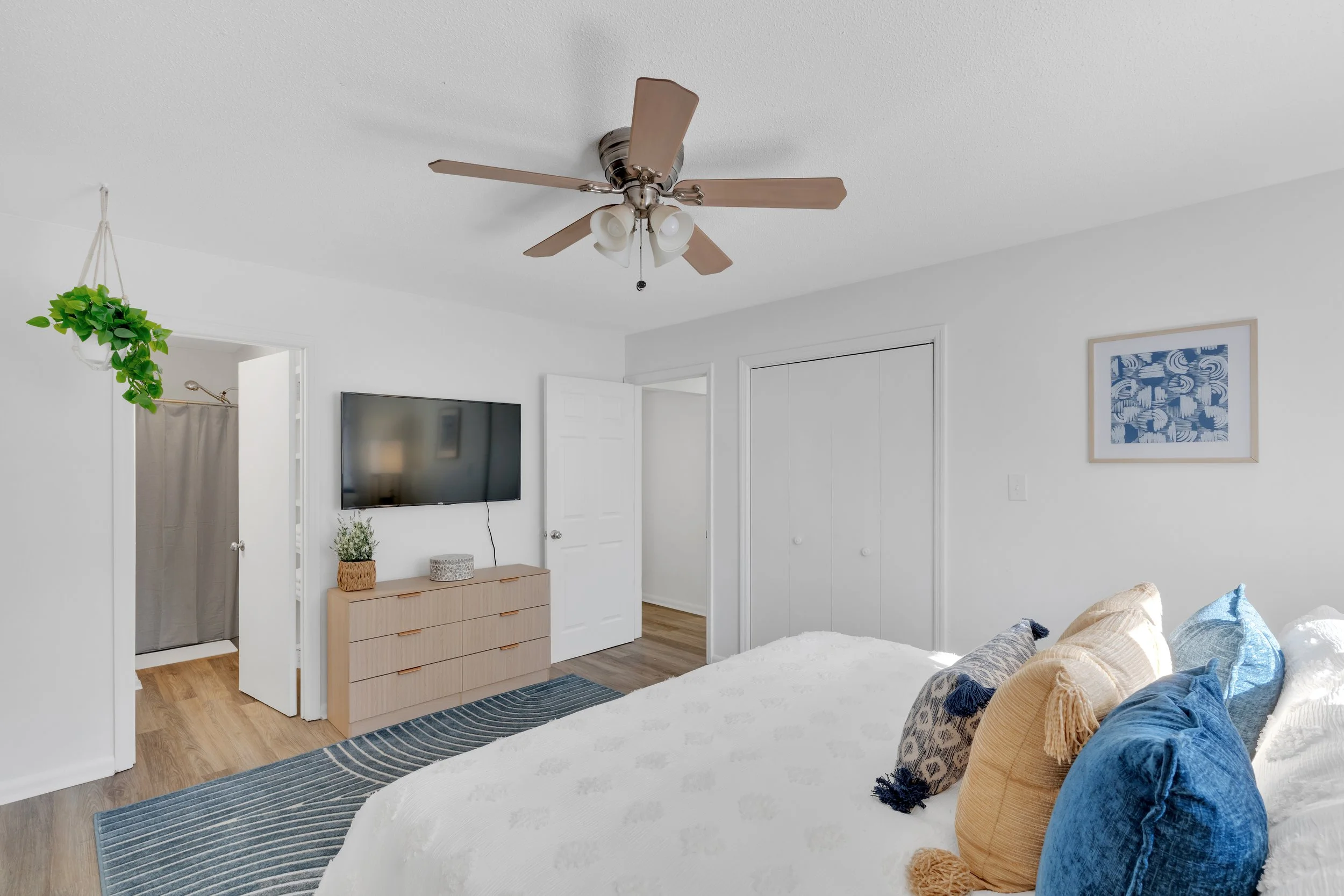 A bedroom with white walls and a ceiling fan. There is a mounted flat-screen TV above a light wood dresser with decorative objects on top. A doorway shows a bathroom with a shower curtain, and a closet with double doors is visible. Part of a bed with