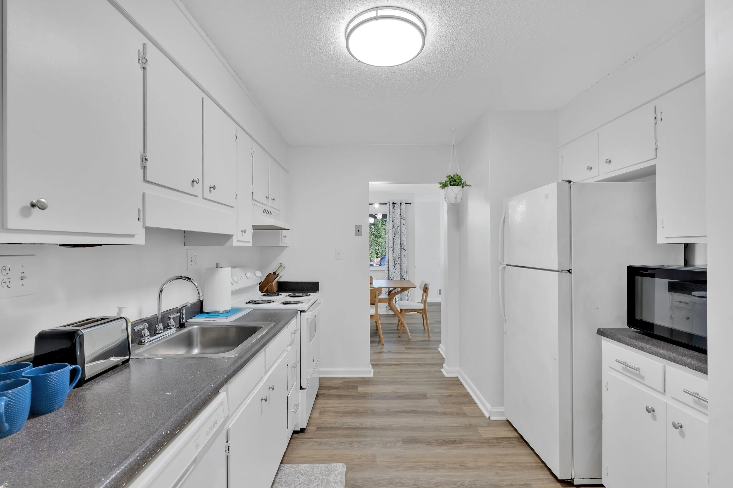 White kitchen with cabinetry and appliances, view toward dining area with table and chairs, wooden flooring, hanging plant, microwave, and living room window visible in the background.