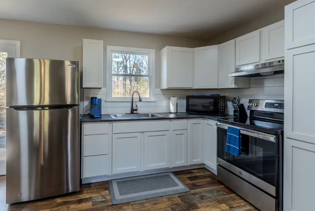 Clean kitchen with white cabinets, stainless steel refrigerator and stove, microwave, black countertop, and hardwood floor.