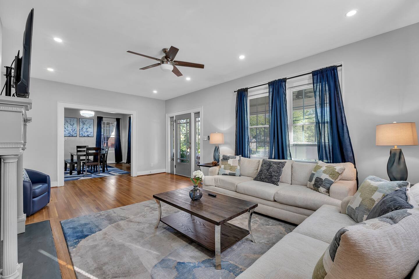 Living room with beige sectional sofa, wooden coffee table, blue curtains, hardwood floor, and adjacent dining area with dark wood table and chairs.