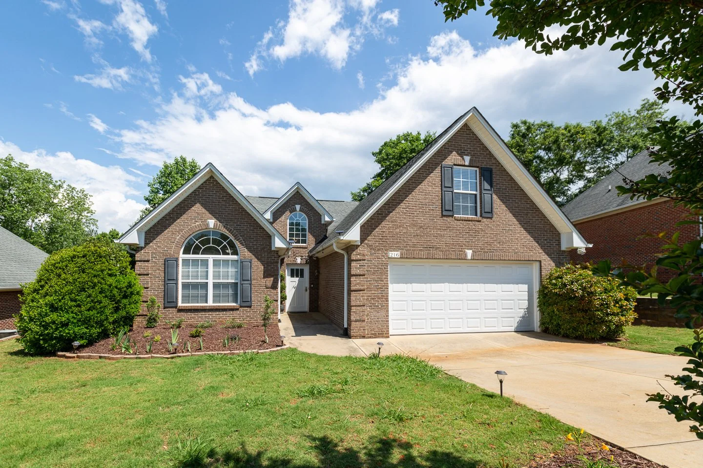 Front view of a brick house with a white garage door and black shutters, surrounded by green shrubs and trees on a sunny day.