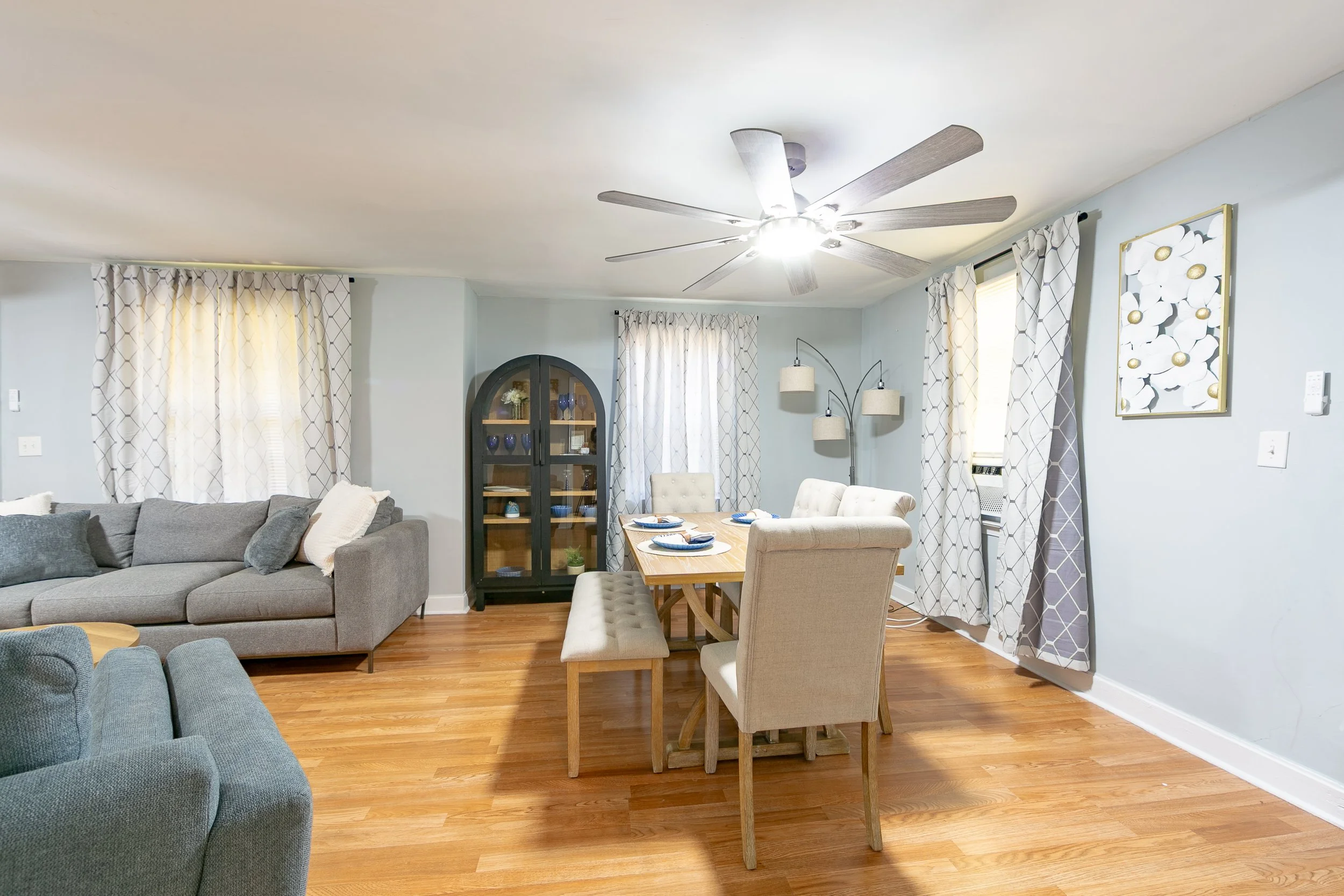 Living room with hardwood floor, light blue walls, a ceiling fan with lights, a gray sofa, a beige dining table with four chairs, a black display cabinet, and curtains with a geometric pattern.