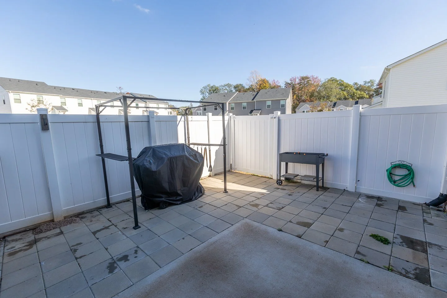 Backyard patio with a white vinyl fence, covered grill, small black grill table, garden hose, and paved ground with a few weeds, under a clear sky.
