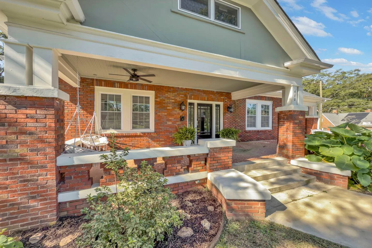 Front porch of a brick house with steps leading up to the black front door, decorative white accents, potted plants, and a hanging swing.