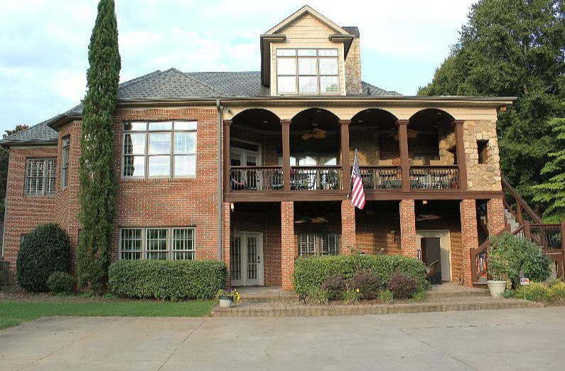 Large two-story brick house with a covered balcony and porch, American flag hanging, surrounded by greenery and landscaping.