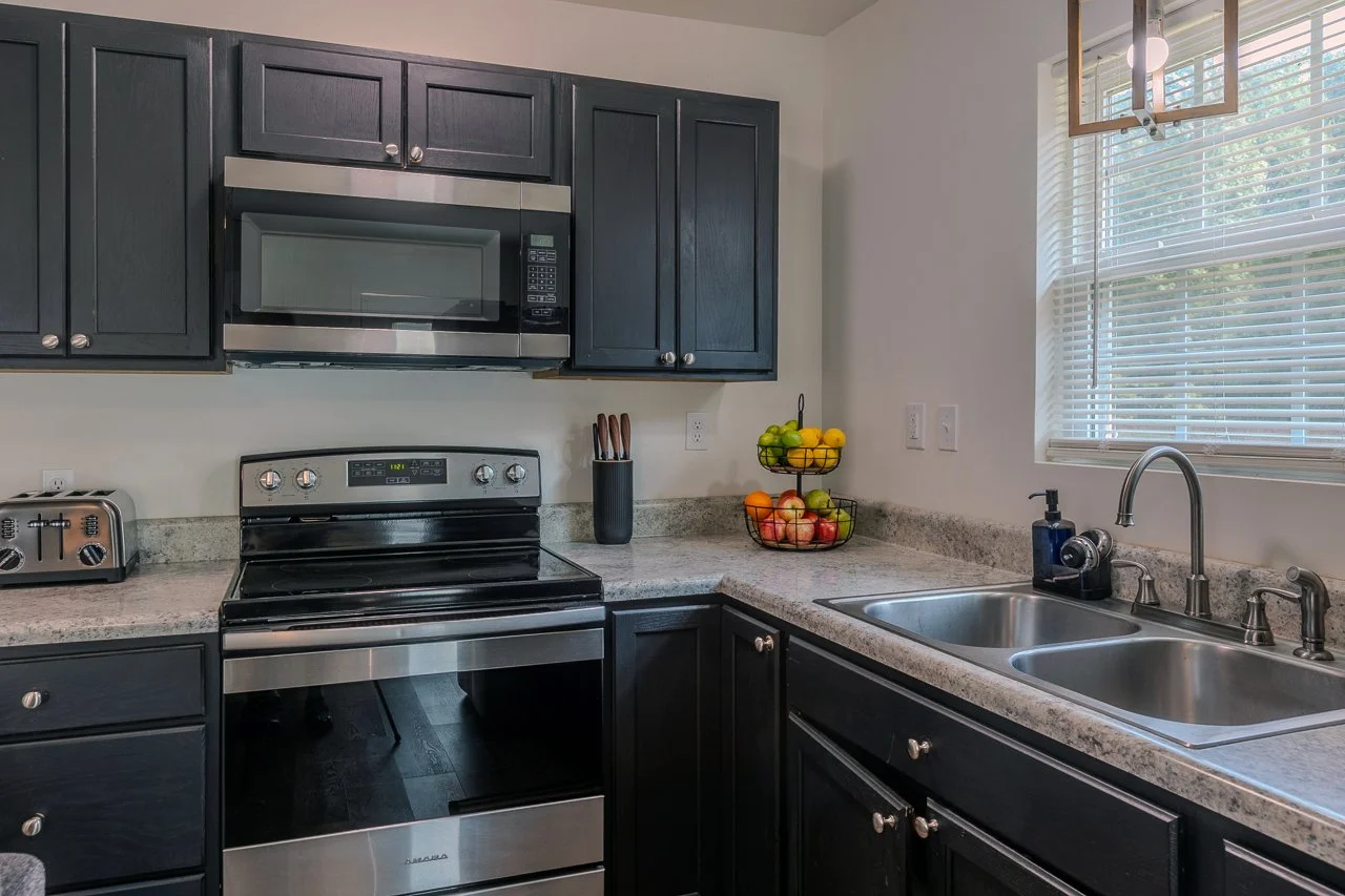 Kitchen with black cabinets, stainless steel microwave and oven, granite countertops, a double sink, window with blinds, and a fruit basket on the counter.