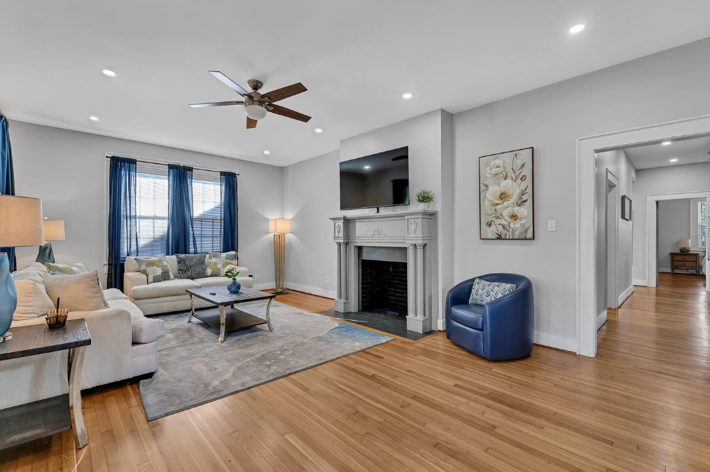 Living room with beige sofas, a coffee table, a fireplace with a mounted TV, blue curtains, a blue armchair, wooden flooring, and artwork on the wall.