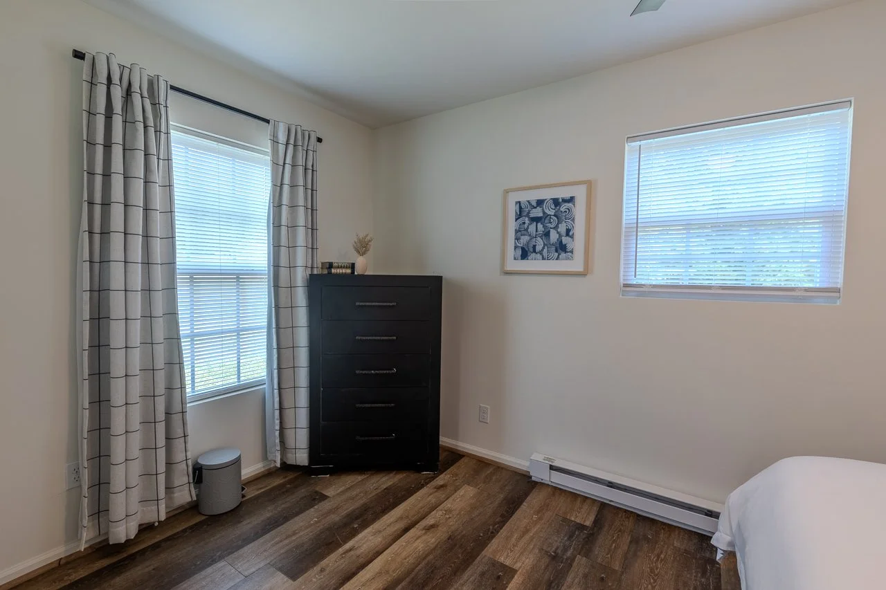 A bedroom with two windows, cream walls, hardwood flooring, and a black dresser. One window has white curtains with a grid pattern, the other has blinds. There is a framed artwork on the wall, and a small trash can near the window.