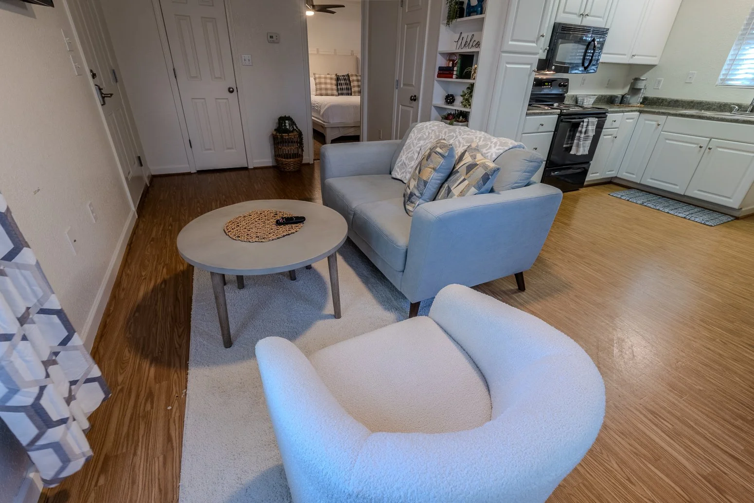 Living room with a white armchair, a light blue sofa with three pillows, a round wooden coffee table with remote controls, a white rug, and a kitchen in the background with white cabinets and stainless steel appliances.