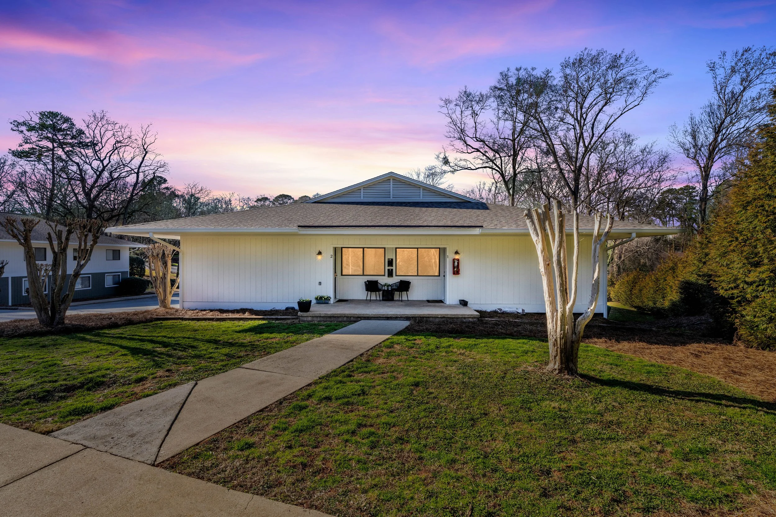 A white house with a small front porch, two black chairs, and a table, set against a colorful sunset sky with pink and purple clouds. The house is surrounded by a green lawn, with three leafless trees in the yard and shrubs on the right side.