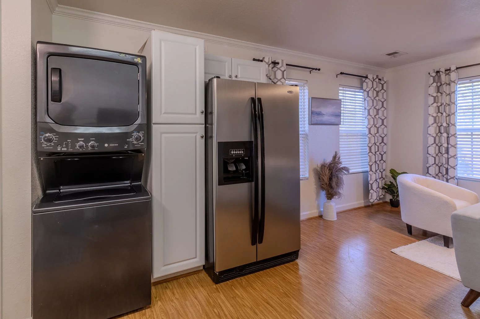 Kitchen area with a stacked washer and dryer, a stainless steel refrigerator, and a living room with white armchair, curtains, and plants.