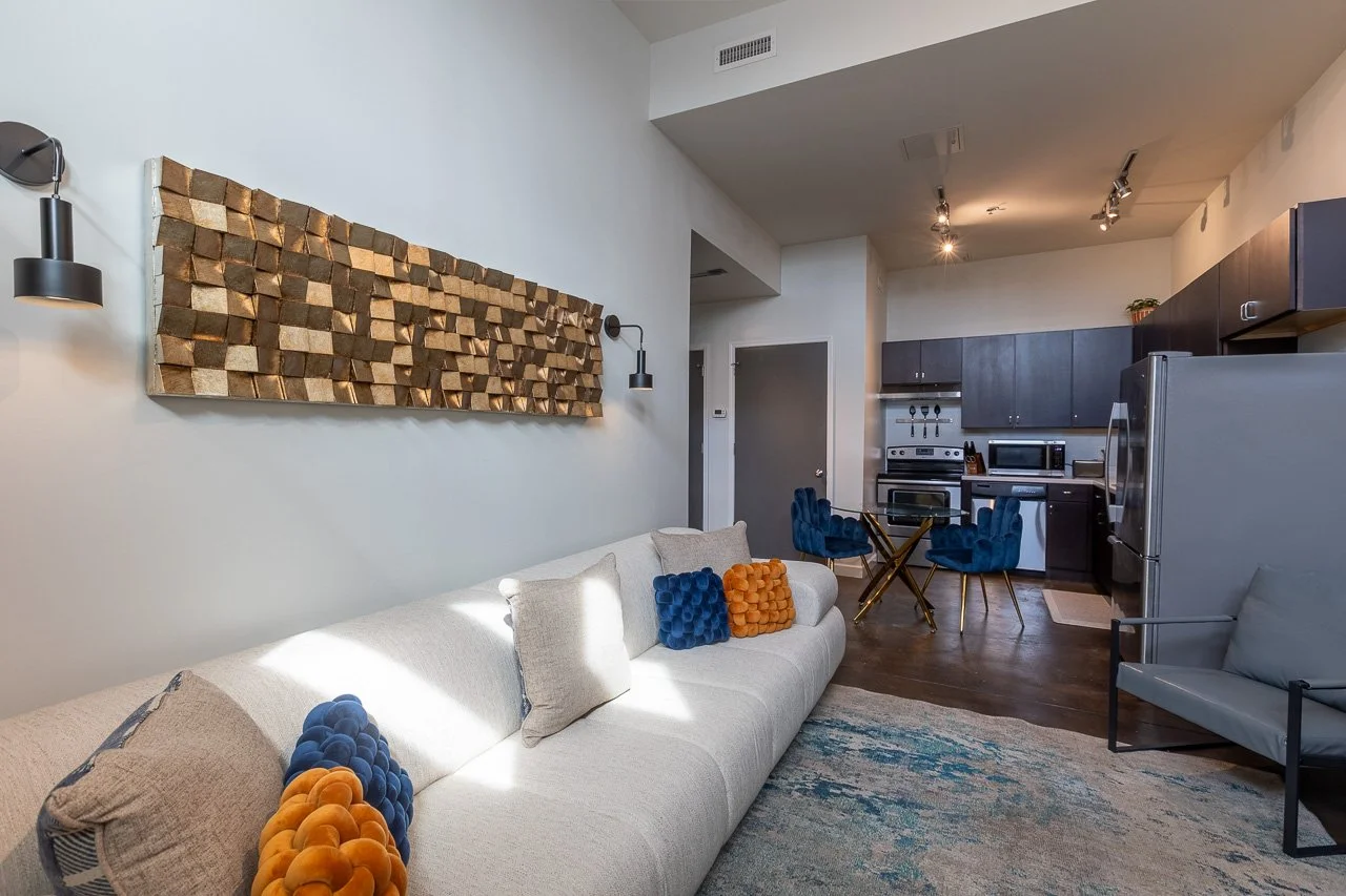Living room with a white sofa and colorful pillows, and a dining area with blue chairs and a small round table, overlooking a kitchen with dark cabinets and stainless steel appliances.