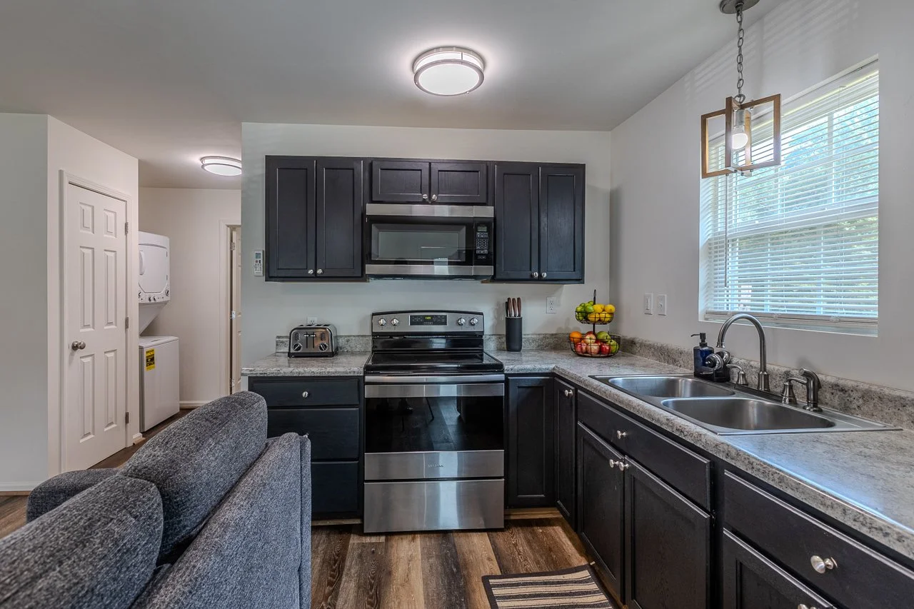 Kitchen with dark cabinets, stainless steel stove and microwave, granite countertops, a double sink, window with blinds, and a small couch in the foreground. A basket of fruit and a toaster are on the counter.