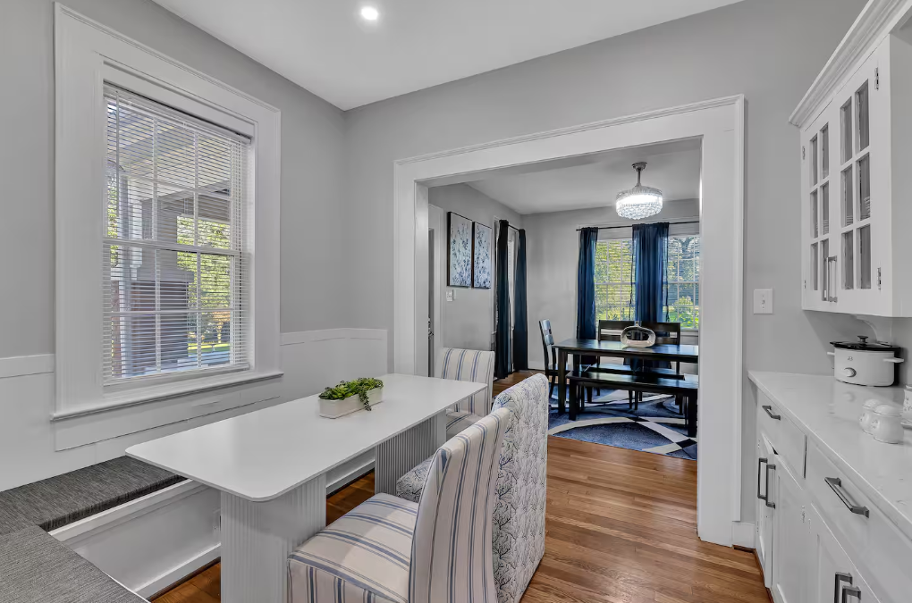 A dining area with a white table, striped chairs, a window with blinds, and a pendant light, leading to a dining room with a table, chairs, dark curtains, and a window showing trees outside.
