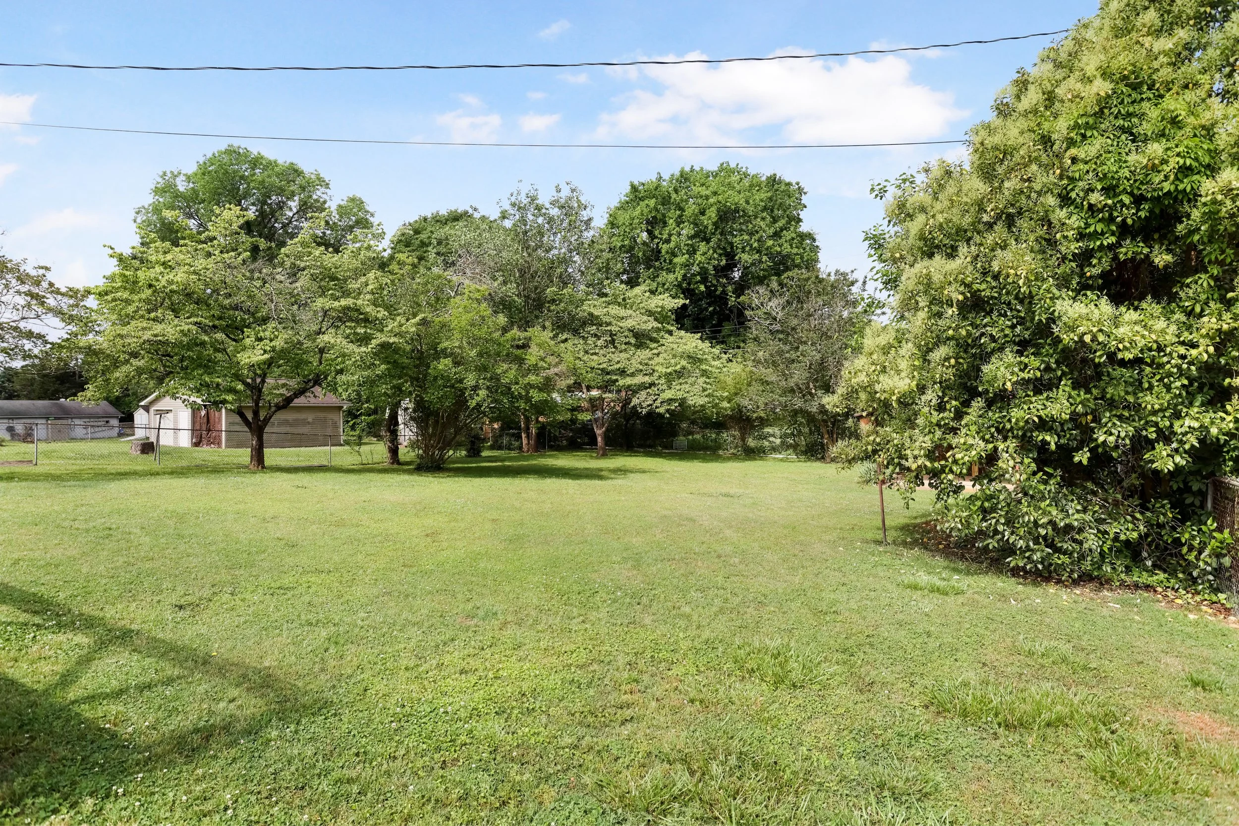 A spacious backyard with a well-maintained grassy lawn, multiple trees providing shade, a small shed in the background, and fence enclosures along the sides. The sky is partly cloudy with some blue visible.