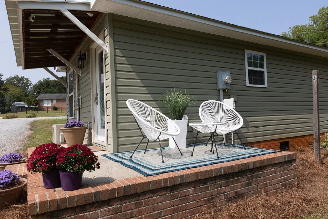 A small porch area with potted purple and red flowers, a striped outdoor rug, two white wire chairs, and a large white planter with green grass-like plants, attached to a gray house with white-framed windows and an electrical meter.