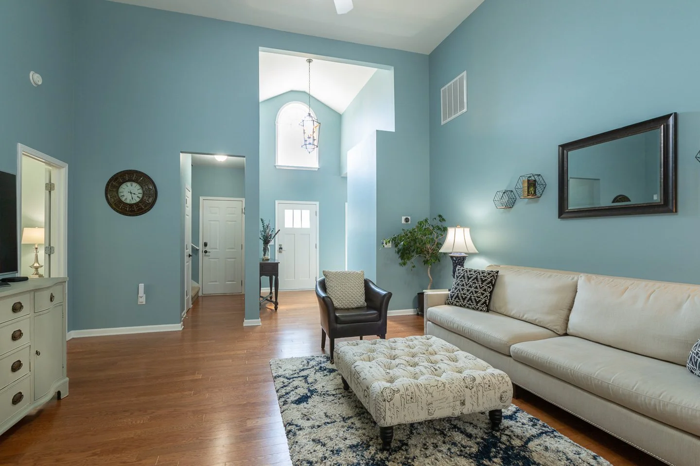 Living room with blue walls, white sofa with black and patterned pillows, black armchair, tufted ottoman, hardwood floors, and wall decor. Entryway visible in background.