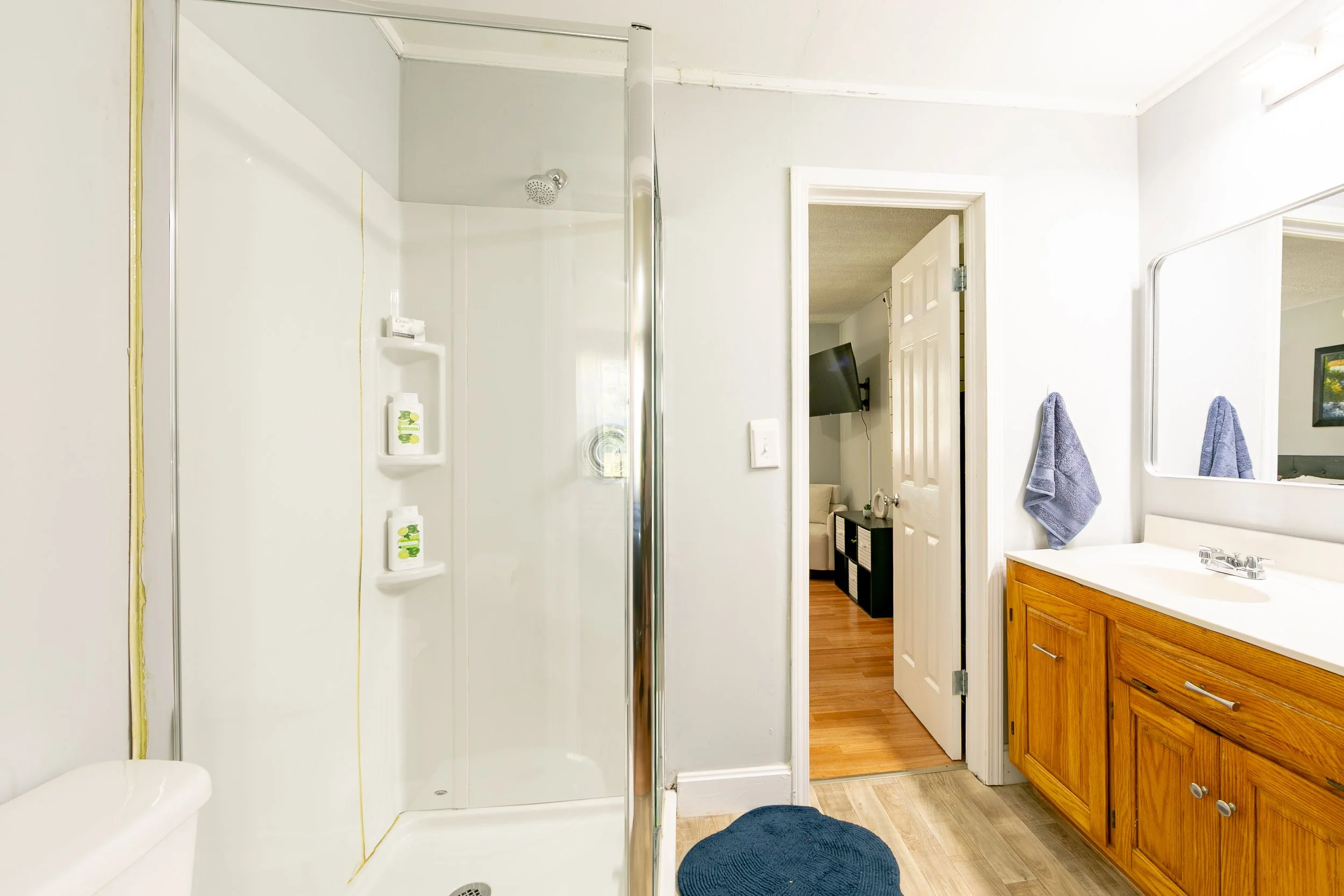 Bathroom with shower, vanity, mirror, and towels, view into adjacent room with TV and wooden floor.