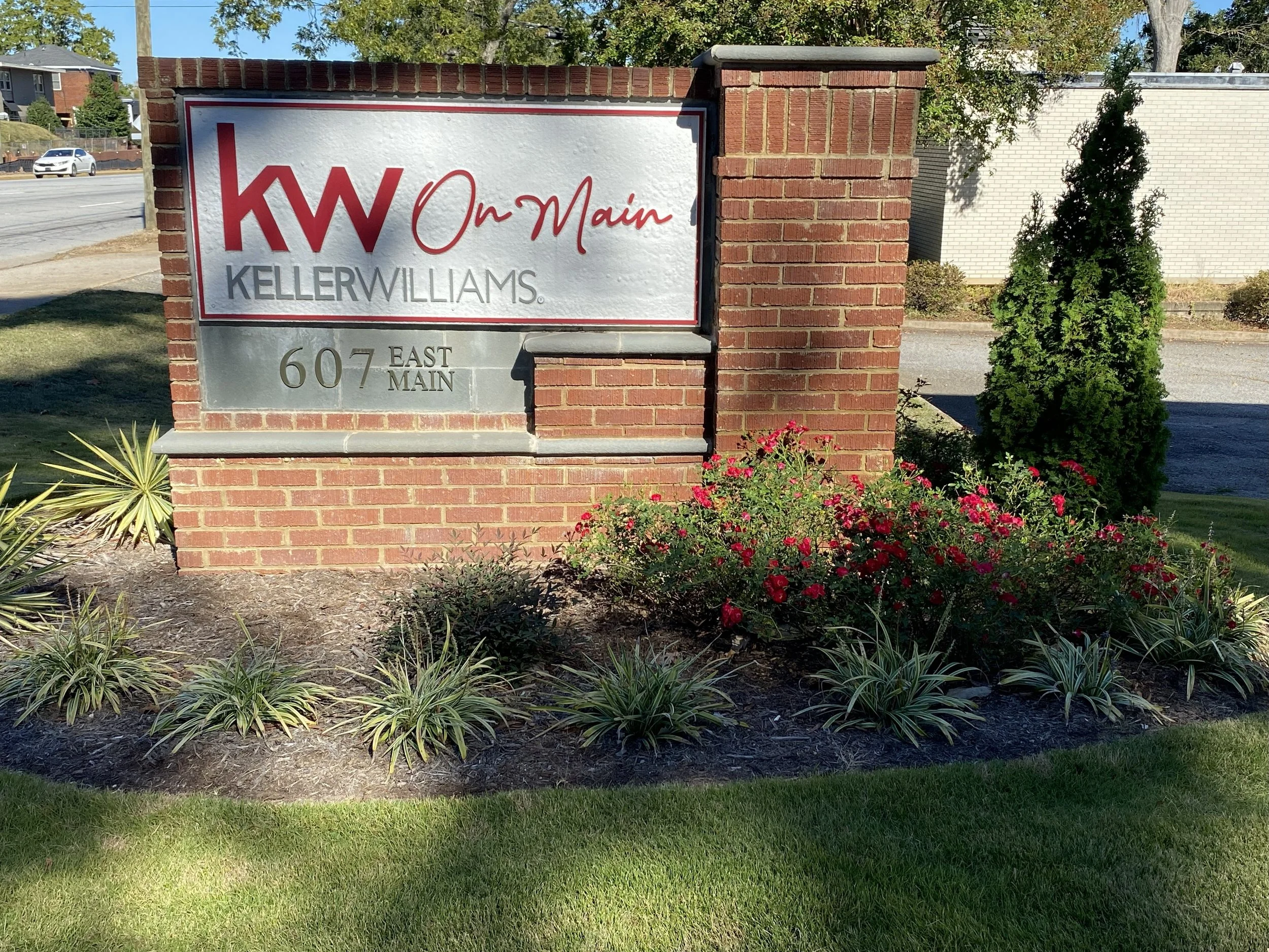 Outdoor sign for Keller Williams real estate agency, with red and gray text, next to flowering plants and a small tree, with a brick and white wall background.