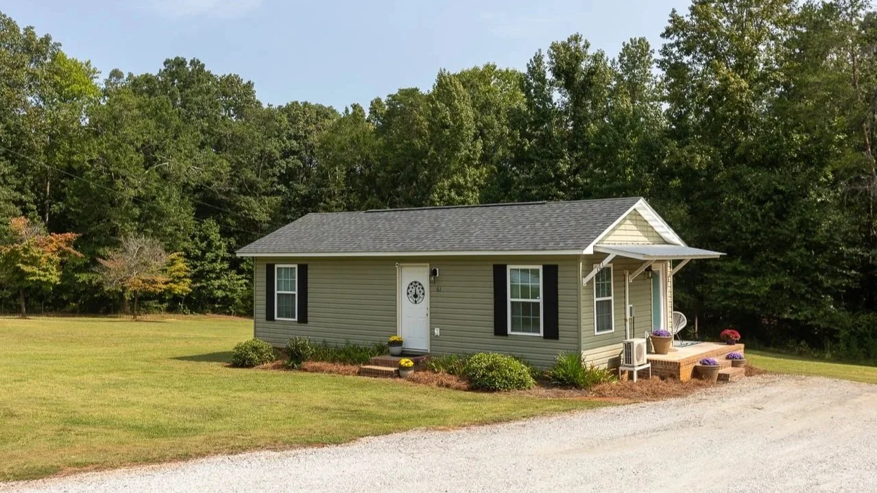 Small house with vinyl siding, black shutters, front porch with plants, surrounded by well-maintained lawn and trees in the background.
