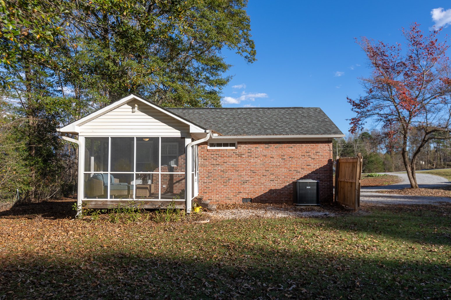 Backyard view of a house with a screened porch, brick and white siding exterior, surrounded by trees with autumn leaves, a grassy yard, and a gravel driveway.