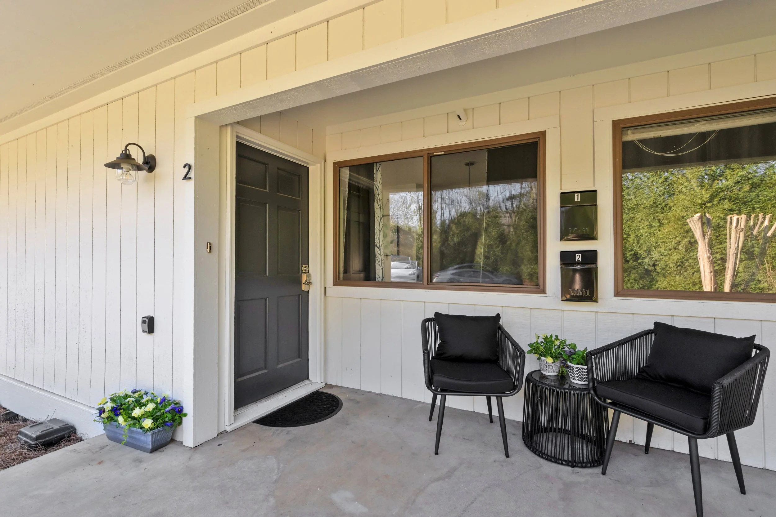 Front porch of a house with black chairs, a small table with potted plants, mailboxes, door, and window with a reflection of trees.