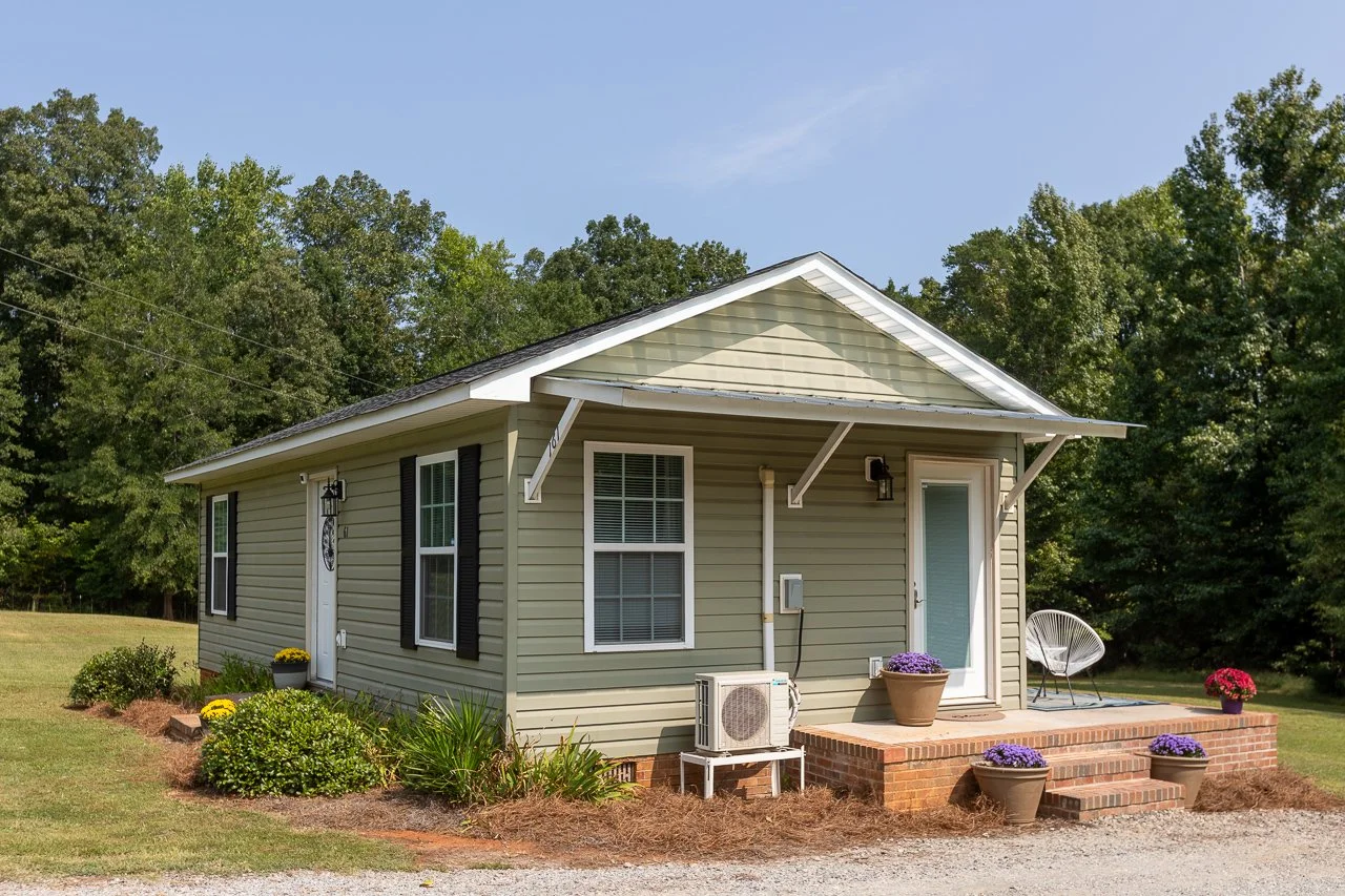 A small, single-story house with beige siding, black shutters, and a gabled roof. The house has a brick porch with steps, potted flowers, and an outdoor chair. There are green bushes and flowers around the porch, and a grassy lawn with trees in the b
