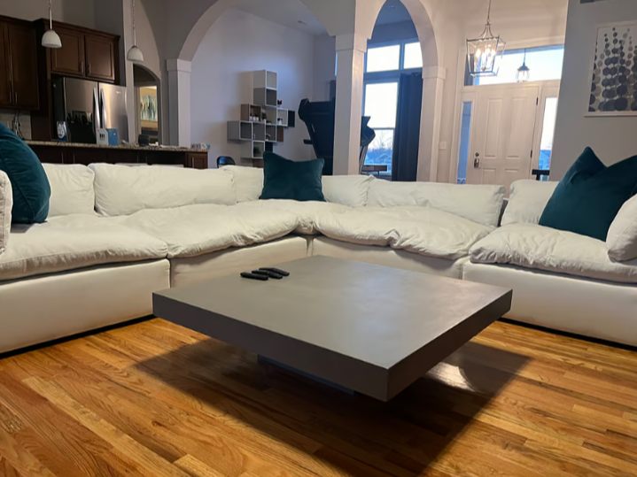Living room with a white sectional sofa, dark teal pillows, a wooden floor, a gray coffee table with three remote controls, and large windows with a front door nearby.