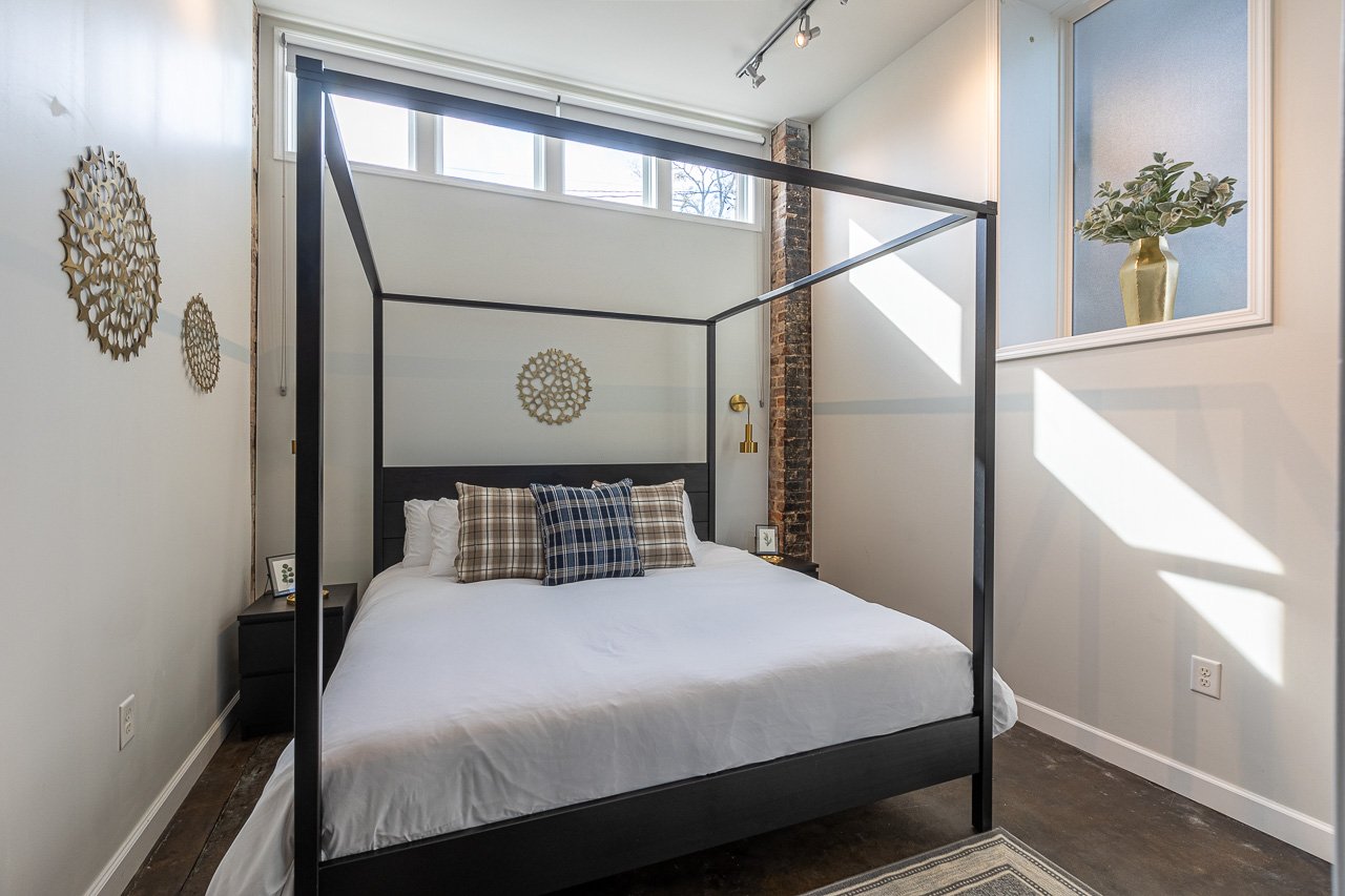 Modern bedroom with a black canopy bed, white bedding, plaid pillows, brass wall sconces, and decor, sunlight coming through high windows.