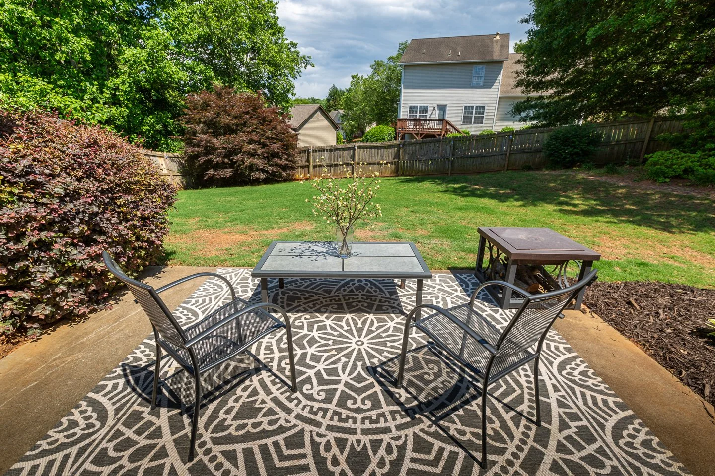 Backyard patio with a patterned outdoor rug, a gray table with a flower vase, two black metal chairs, and a small wooden fire pit table surrounded by green trees and grass.