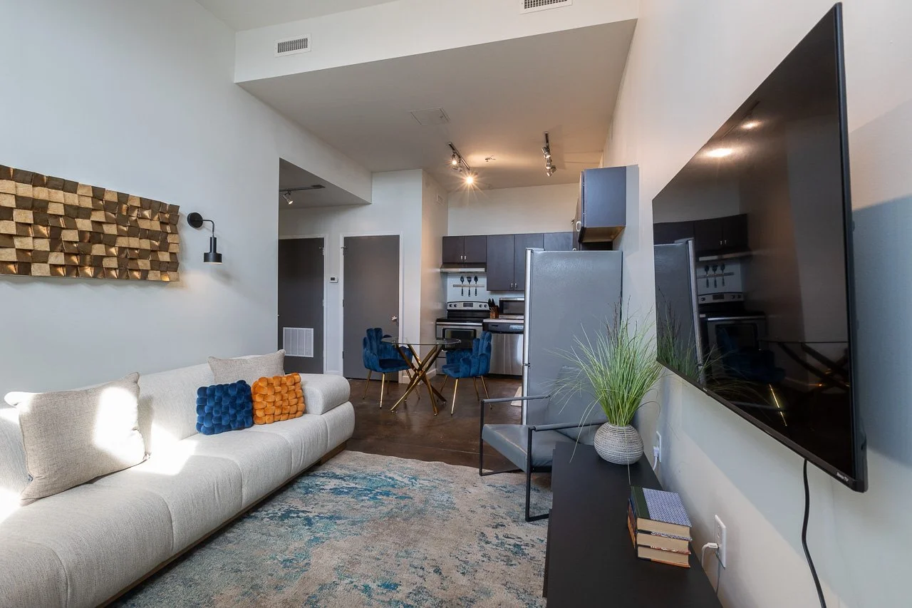 Living room with a white sofa featuring two decorative pillows, flat-screen TV on the wall, potted plant, and an open kitchen with dark cabinets and a dining table with blue chairs.