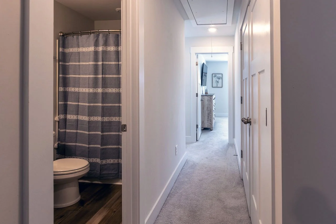A hallway in a residential home with a bathroom entrance to the left and a doorway at the end leading to a bright room with a TV and framed artwork on the wall.