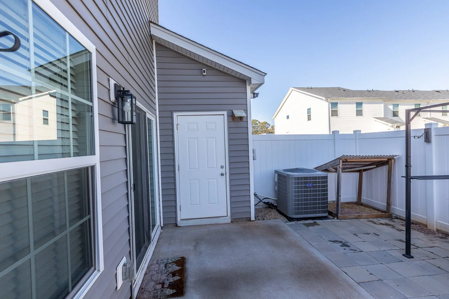 Backyard patio area with siding house, sliding glass door, outdoor light fixture, white door, air conditioning unit, small wooden shelter, and paver stones on ground, enclosed by white fence.