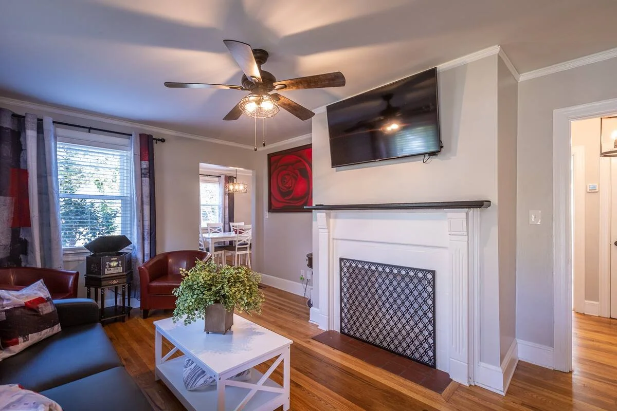 Living room with a ceiling fan, a wall-mounted TV, a white fireplace with decorative trim, hardwood floor, armchairs, a coffee table with a plant, and windows with curtains.