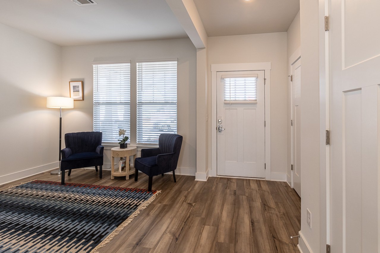 Living room corner with two navy blue armchairs, a small beige side table with a potted plant, window blinds, a floor lamp, and a multicolored rug, with white walls and hardwood floors.