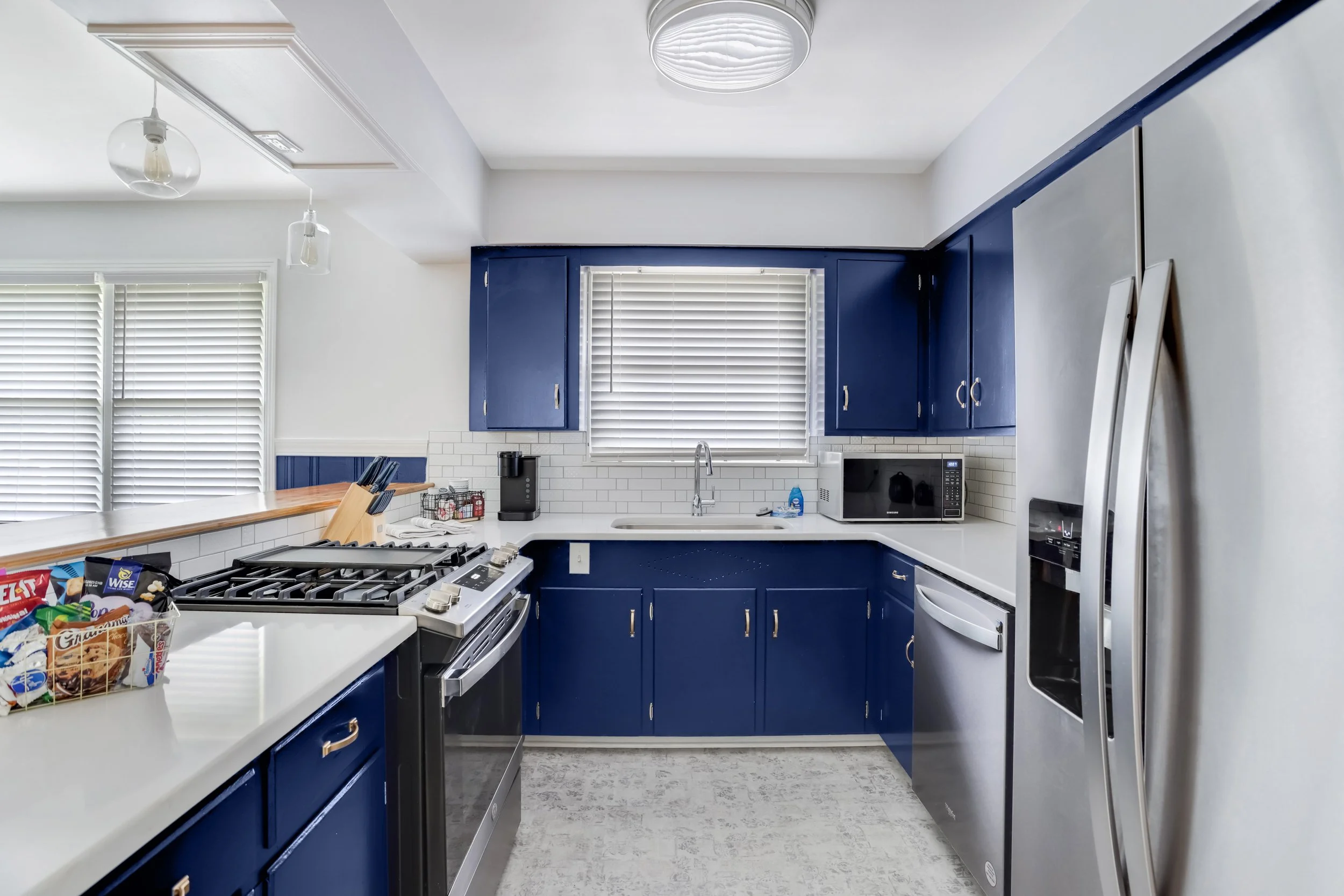 Modern kitchen with blue cabinets, stainless steel appliances, white countertops, and white subway tile backsplash. Contains a gas stove, microwave, refrigerator, coffee maker, and various kitchen items.