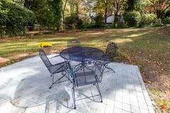 Outdoor patio with a round metal table and four matching chairs on a tiled surface, surrounded by grass, trees, and a yellow fire pit in the background.