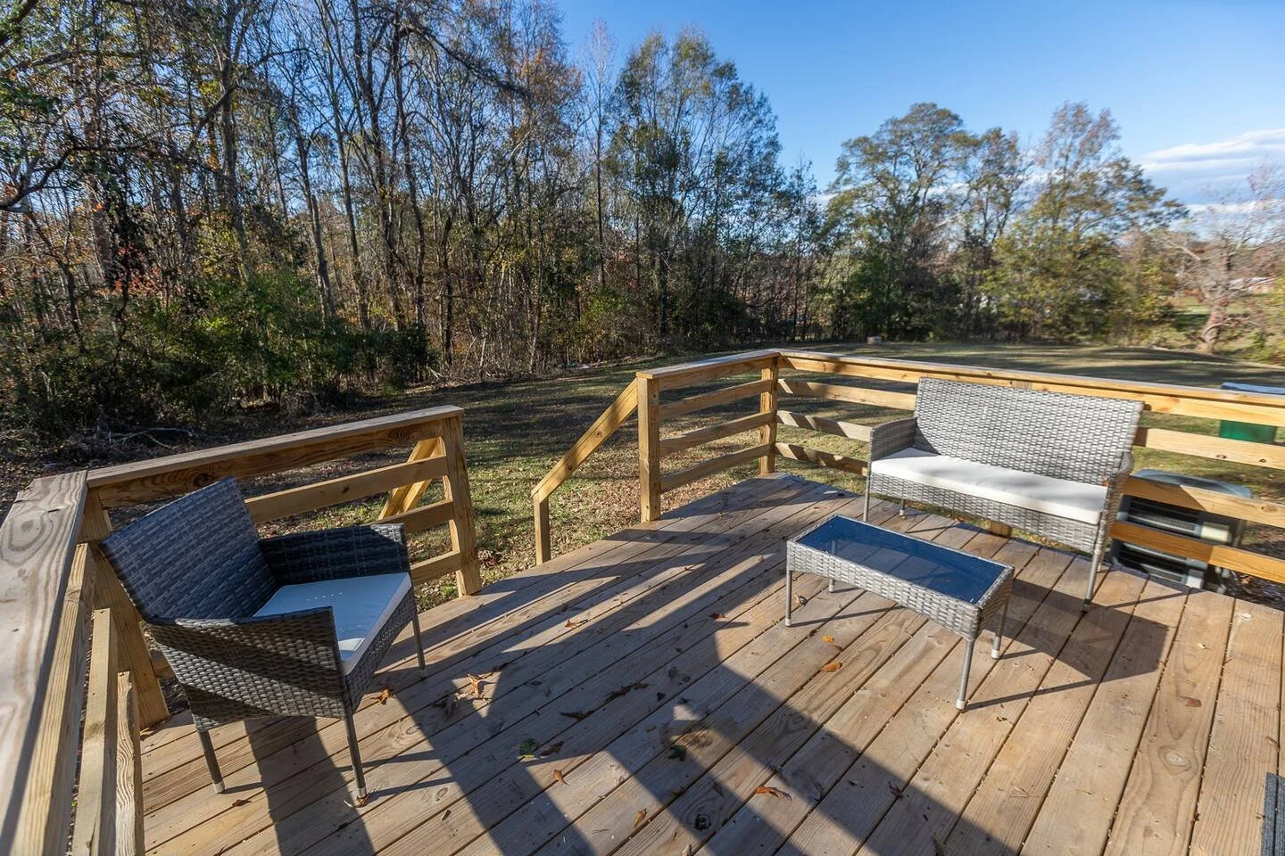 View from a wooden deck with patio furniture including two chairs, a loveseat, and a small glass-top table, overlooking a backyard with grass and trees under a clear blue sky.