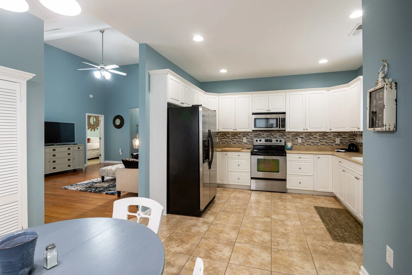 Kitchen with white cabinets, stainless steel refrigerator, stove, microwave, beige tile floor, blue walls, and a round blue table with white chairs in the foreground.