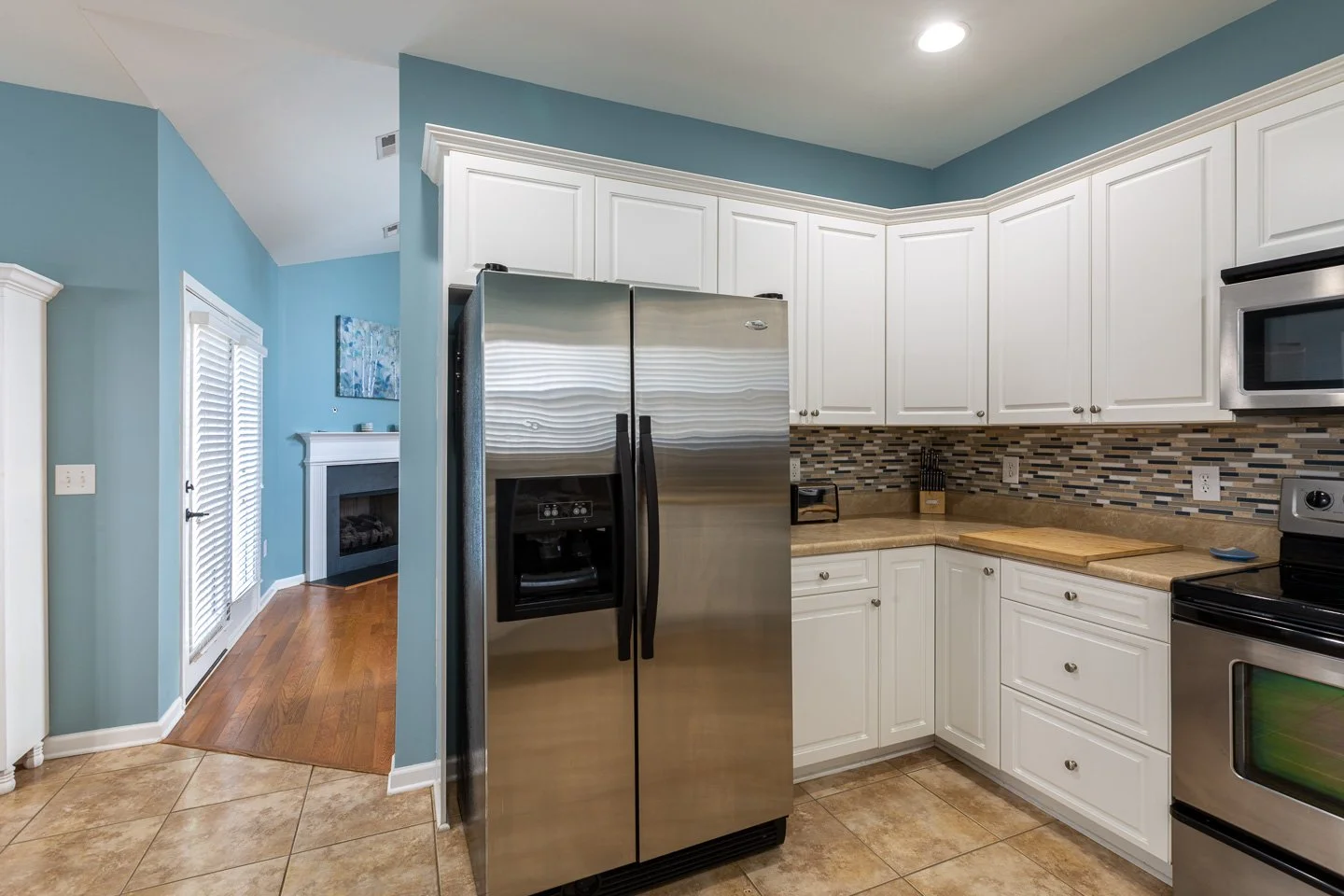 A modern kitchen with white cabinets, stainless steel appliances, and a teal wall, overlooking a living room with hardwood flooring and a fireplace.