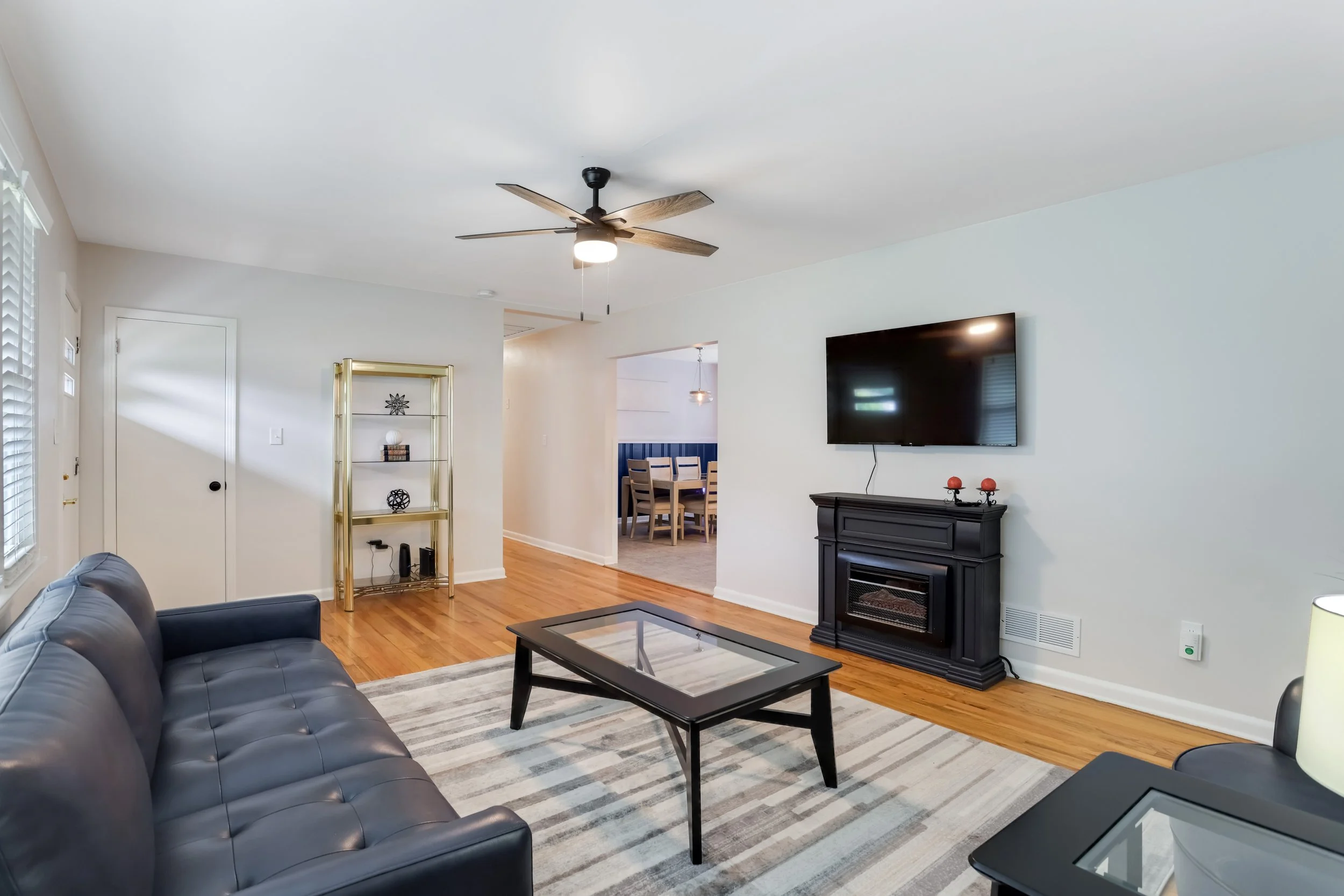 Living room with leather sofa, glass-top coffee table, wall-mounted TV, black fireplace, and a gold shelf against white walls and hardwood floors.