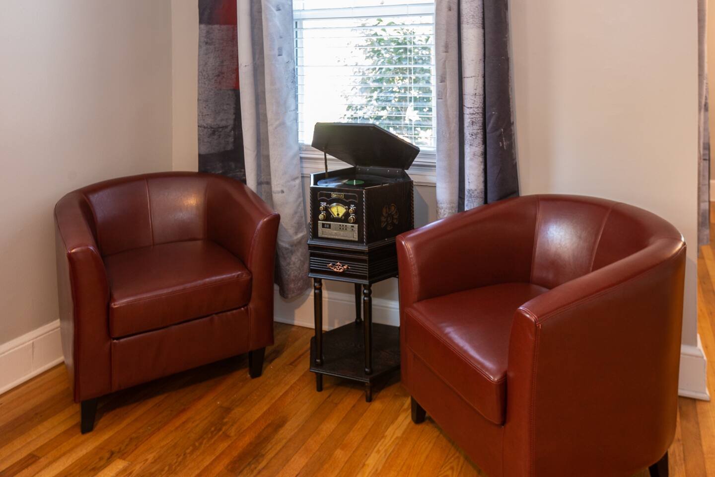 Two brown leather armchairs positioned on either side of a vintage-style black record player on a small black stand in front of a window with partially open blinds and patterned curtains.