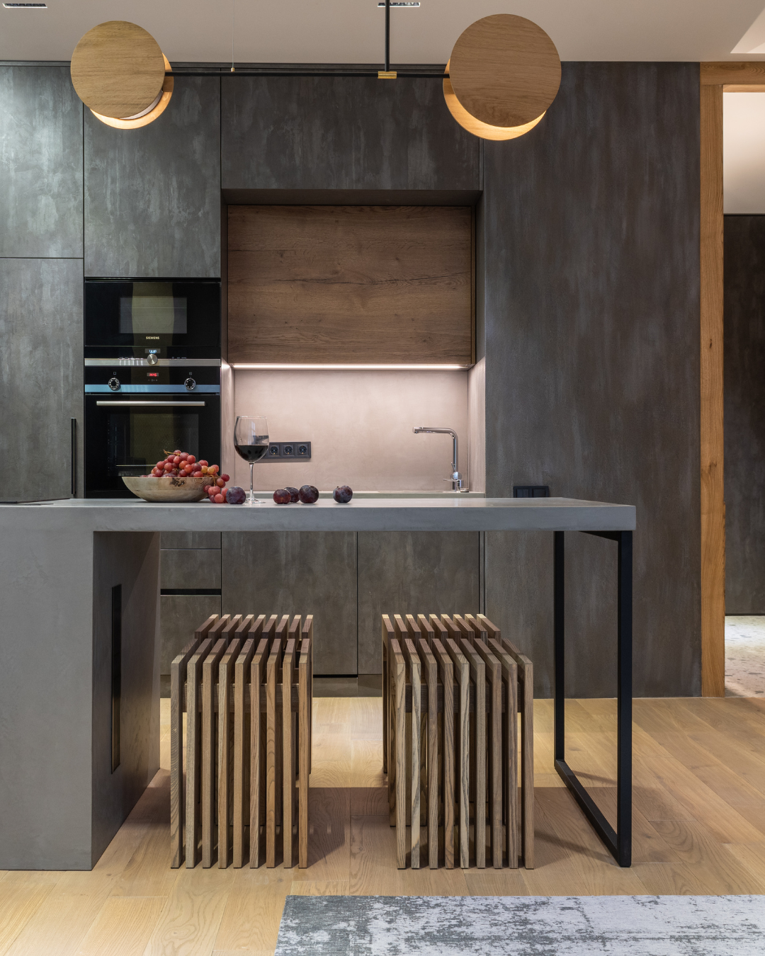 Modern kitchen with concrete and wood cabinetry, a small sink, a glass of red wine, grapes, a bowl, and two wooden stools at a light-colored countertop, with minimalist lighting fixtures hanging from the ceiling.