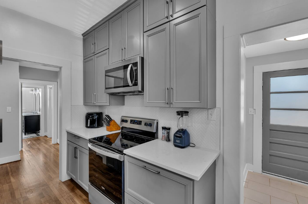 Kitchen with gray cabinets, white countertops, stainless steel microwave, stove, and various small appliances including a blender, coffee maker, and spice jars.