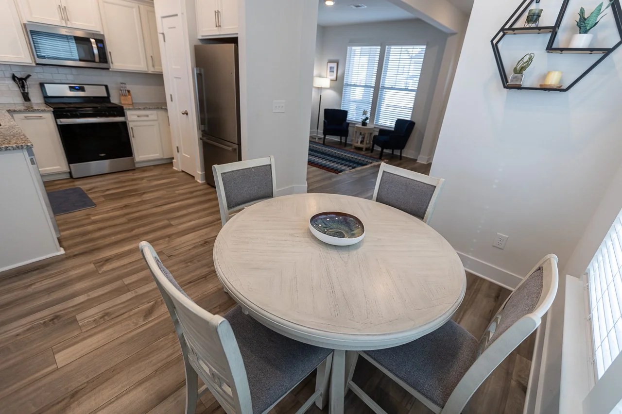 A round dining table with four chairs in a kitchen and dining area. The table has a decorative bowl in the center. The kitchen has white cabinets, a stove, a refrigerator, and granite countertops. There is a seating area with two chairs and windows i