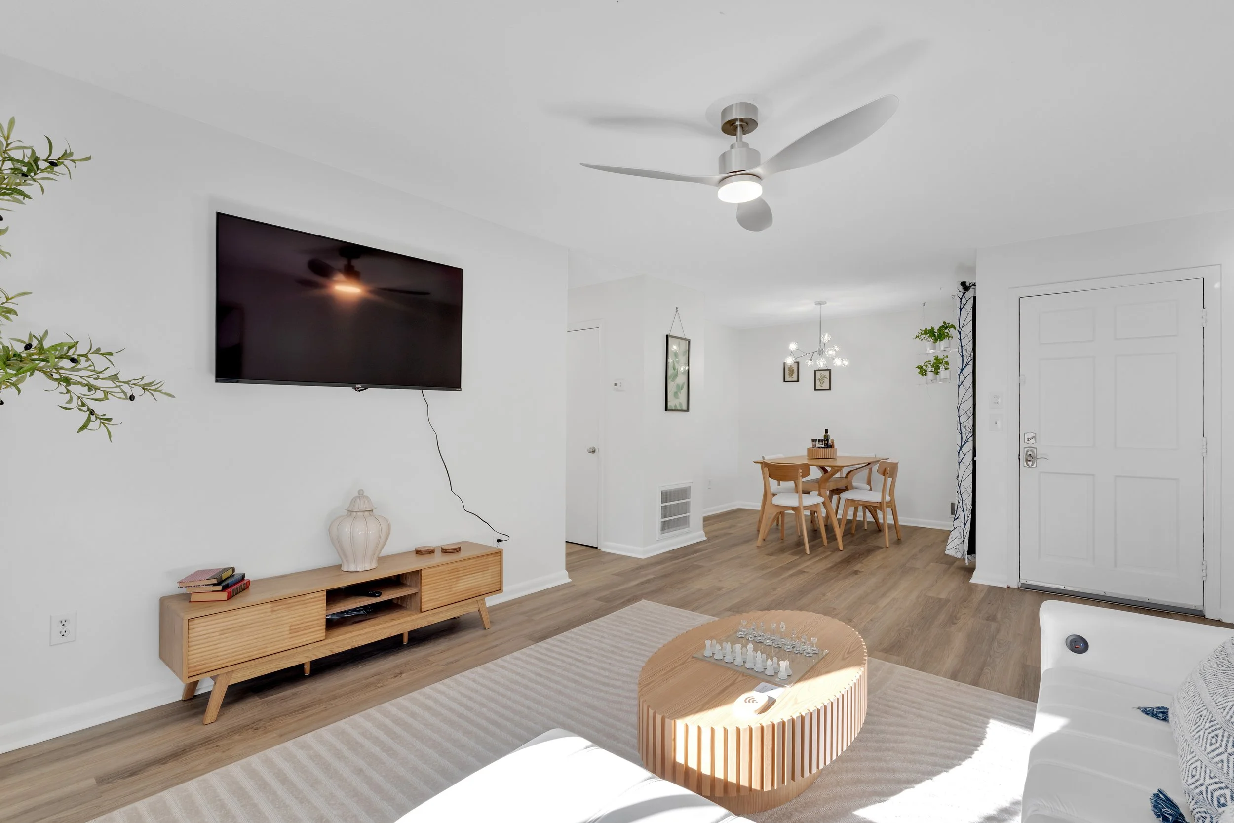 Living room with wall-mounted TV, wooden TV stand, light-colored sofa, round wooden coffee table, dining area with four chairs, a chandelier, and plants, with white walls and wood flooring.