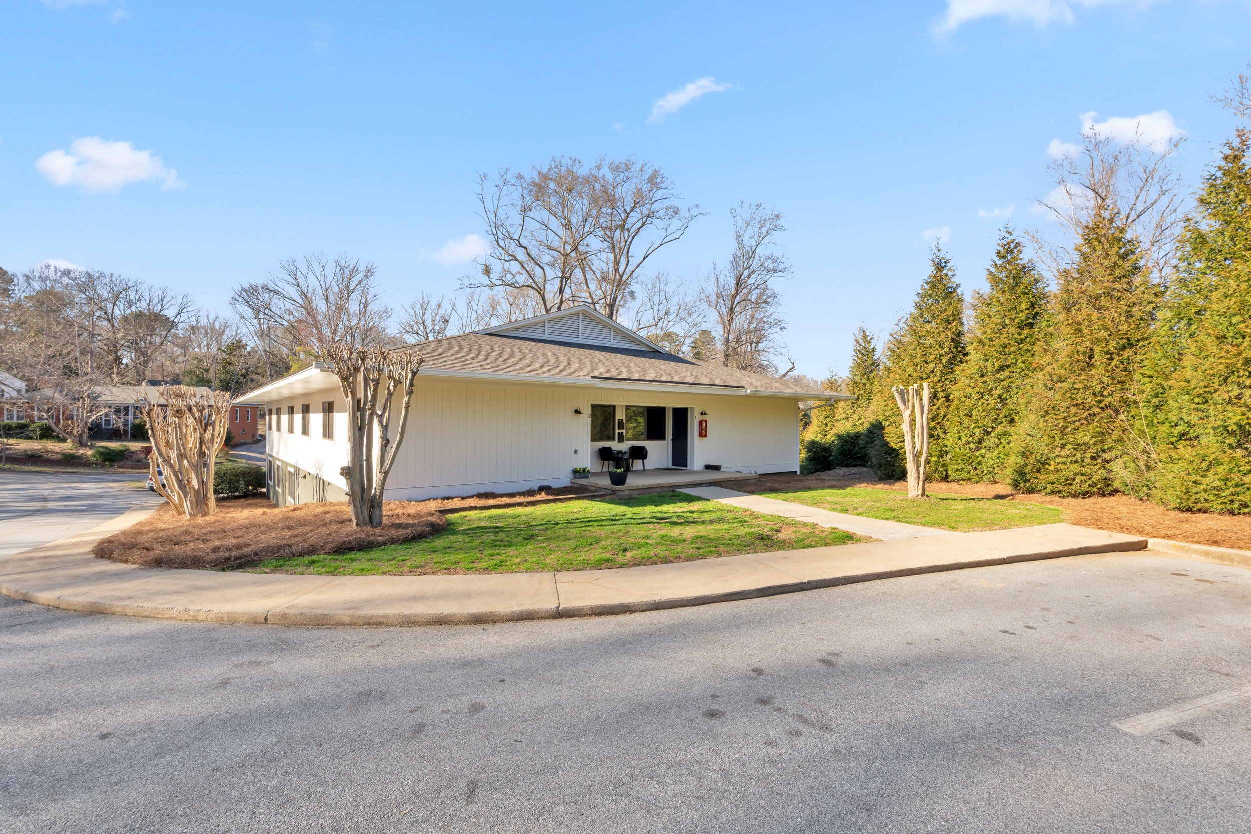 A white single-story building with a sloped roof, front porch, and small potted plants, surrounded by trees with some leafless and others evergreen, under a blue sky with a few clouds.