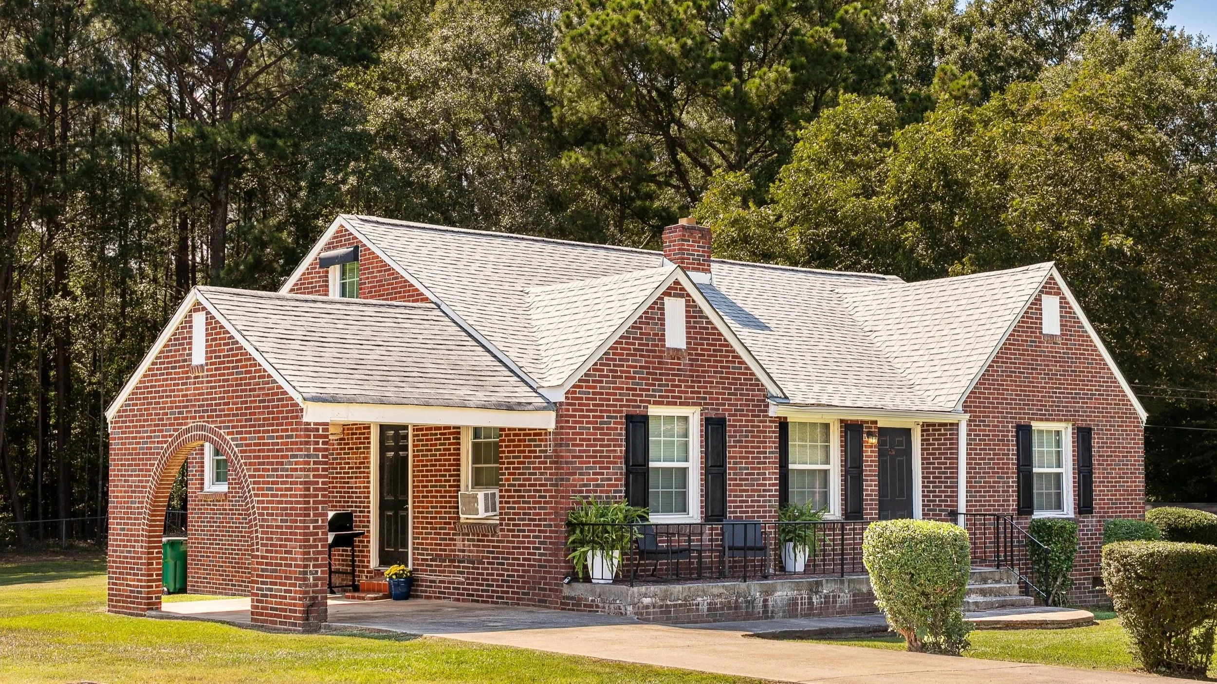 A brick house with a white shingle roof, black shutters, and a small front porch with black railing. The house has a brick chimney and several windows, with two potted plants on the porch. The lawn is green and well-maintained, with bushes and trees in the background.